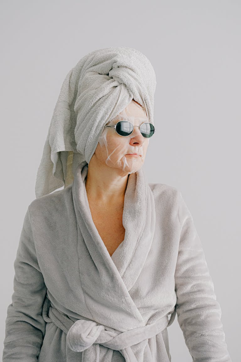 Aged female in gray bathrobe and with towel on head standing on gray background and looking away in sunglasses with sheet mask on face- symbolizing Entering Your 30s and 40s